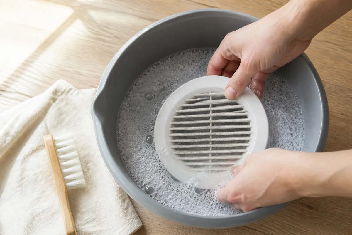 Mains nettoyant une bouche d’extraction blanche démontée dans une bassine d’eau savonneuse
